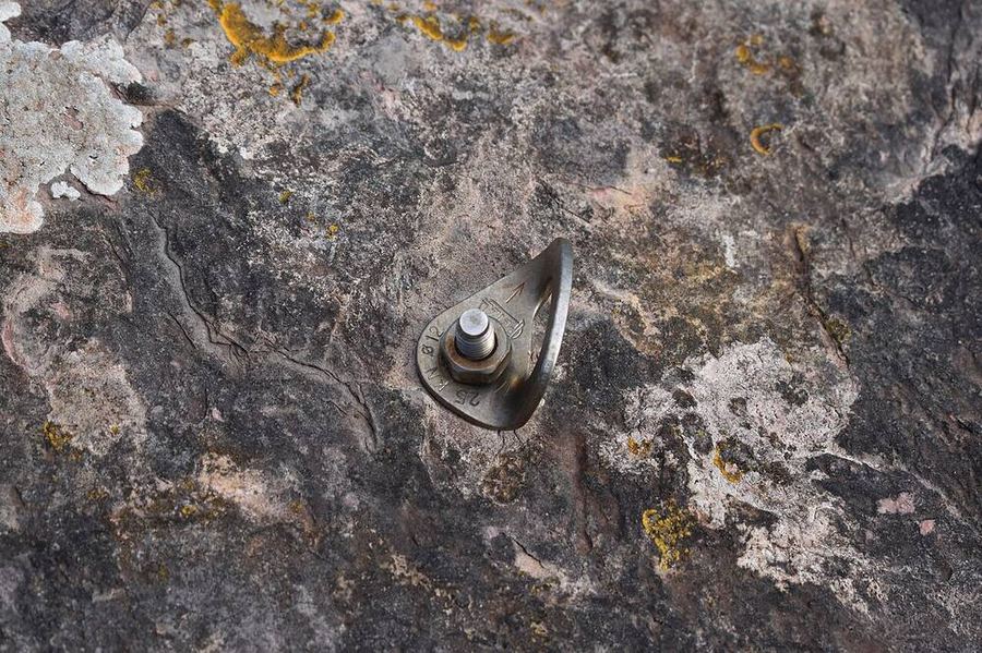 Plaquette inox en forme de goutte, marquée « Ø 12 » et « 25 kN », serrée par un écrou sur un goujon dans une roche couverte de lichens