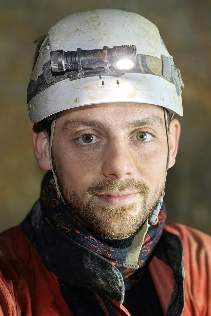 Portrait d'un spéléologue casqué, en combinaison rouge, avec le col noir d'une sous-combinaison et un tour de cou