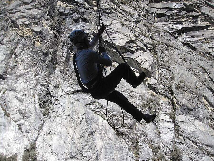 Grimpeur casqué pendu sous un surplomb, une main sur un nœud de cordelette serré sur la corde, un pied dans une boucle